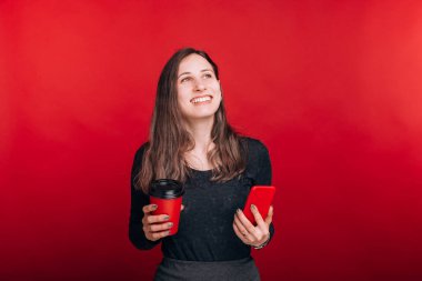 Genç gülümseyen kadın kahvesinin tadını çıkarıyor, telefonunu tutuyor, yukarı bakıyor ve arka planda bir şeyin hayalini kuruyor..