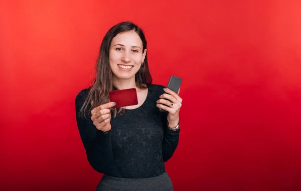 La banque mobile est géniale. Joyeux jeune femme tenant une carte de crédit et son téléphone sur fond rouge — Photo