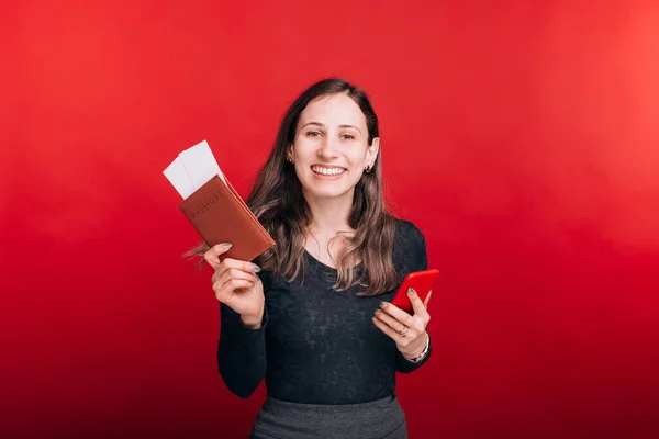 Nadšená mladá žena ukazuje svůj pas a nějaké vstupenky v něm a drží svůj telefon na červeném pozadí — Stock fotografie