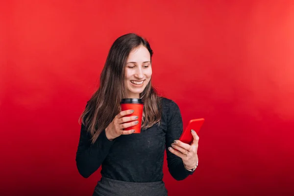 Joven mujer sonriente está mirando su teléfono y sosteniendo una taza de papel para ir sobre fondo rojo — Foto de Stock