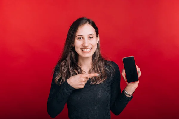 Olha para esta oferta. Mulher sorridente está apontando para um telefone em branco no fundo vermelho — Fotografia de Stock