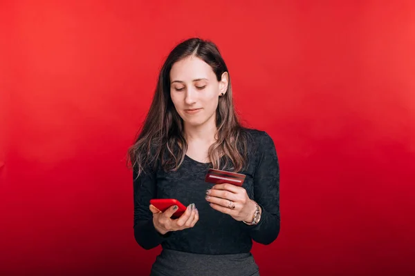 Güzel genç bir kadın telefonuna bakıyor, internetten bir şeyler alıyor ve kırmızı arka planda kredi kartıyla ödeme yapıyor. — Stok fotoğraf
