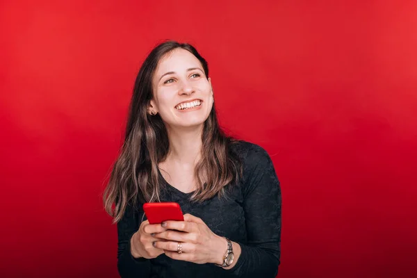 Jó hírek. Mosolygó fiatal nő felnéz, miközben ő tartja az okos telefon piros háttér — Stock Fotó