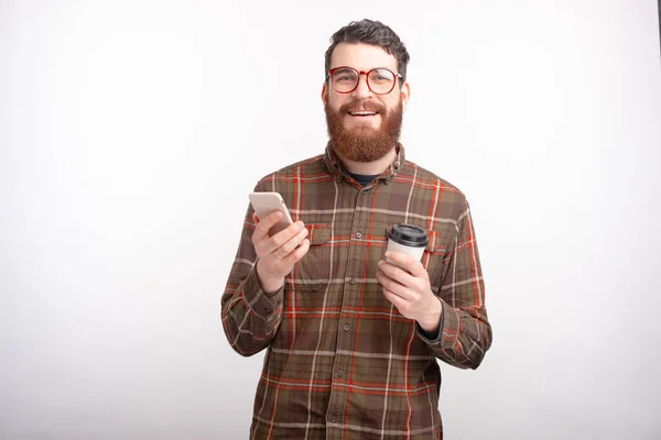 Joven alegre con barba sosteniendo teléfono inteligente y taza de café para llevar — Foto de Stock