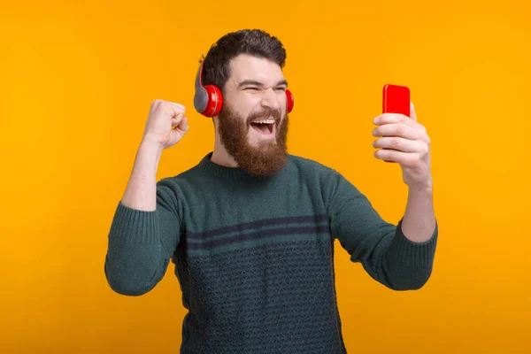 Escuchar en línea es una gran cosa. Barbudo hombre está viendo algo en su teléfono, usando auriculares y haciendo un gesto ganador en el fondo amarillo —  Fotos de Stock