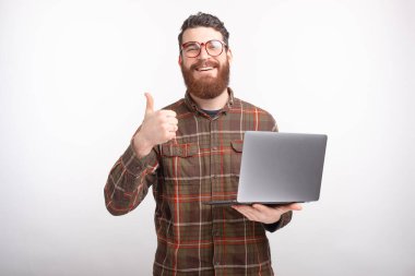 Yakışıklı genç sakallı adam dizüstü bilgisayarını beyaz arka planda tutarken başparmağını kaldırıyor ya da hareket ediyor.