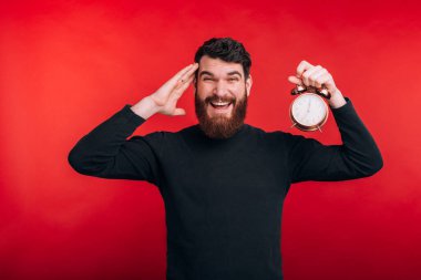 Şaşırmış sakallı adam kırmızı arka planda duruyor ve çalar saati tutuyor.