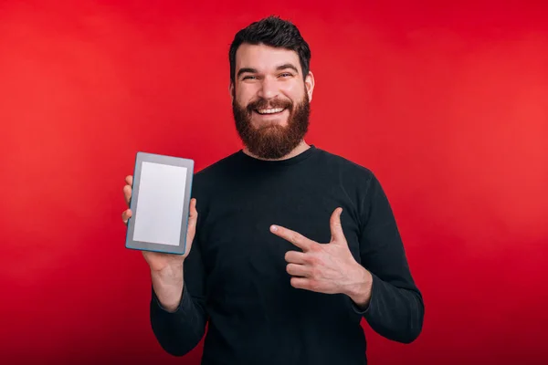 Hombre barbudo feliz apuntando a la pantalla de la tableta sobre fondo rojo — Foto de Stock