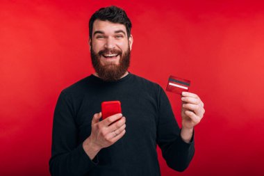 Sakallı, akıllı telefon tutan ve kredi kartı gösteren neşeli mutlu bir adam.