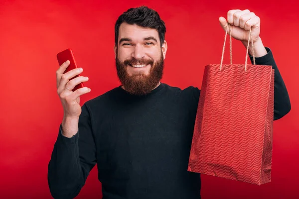 Jovem feliz com barba segurando smartphone e saco de compras, compras on-line — Fotografia de Stock