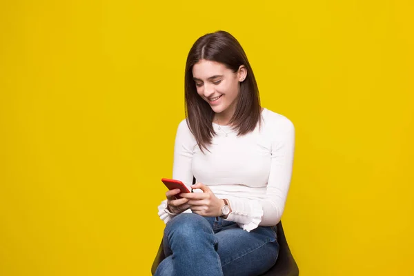 Menina bonita jovem sentado em uma cadeira e segurando telefone celular em mãos, compras on-line no fundo amarelo . — Fotografia de Stock
