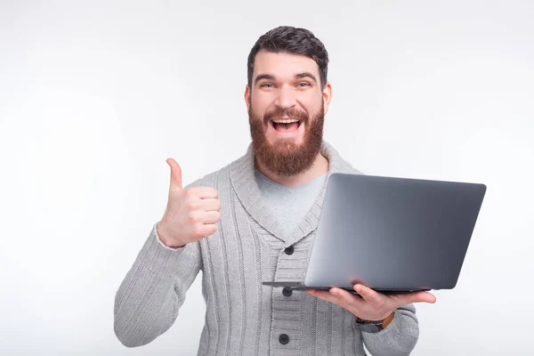 Erstaunter Mann mit Bart steht vor weißem Hintergrund, während er Laptop hält und Daumen nach oben zeigt — Stockfoto