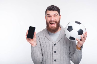 Akıllı telefon ve futbol topunu beyaz arka planda tutan genç, şaşkın bir adam.