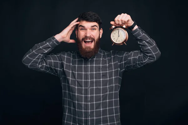 Joven con barba sosteniendo reloj despertador sobre backgorund negro — Foto de Stock
