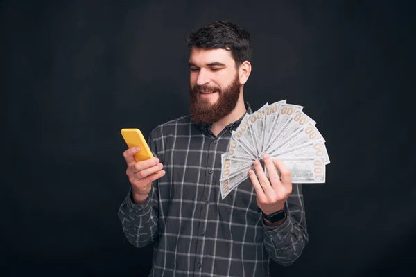 一个留着胡子的年轻人用智能手机拿着钱 — 图库照片