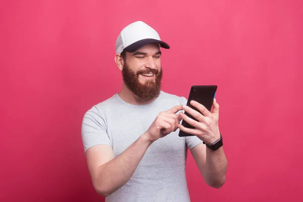 Homme gai avec la barbe souriant et en utilisant sa tablette sur fond rose — Photo
