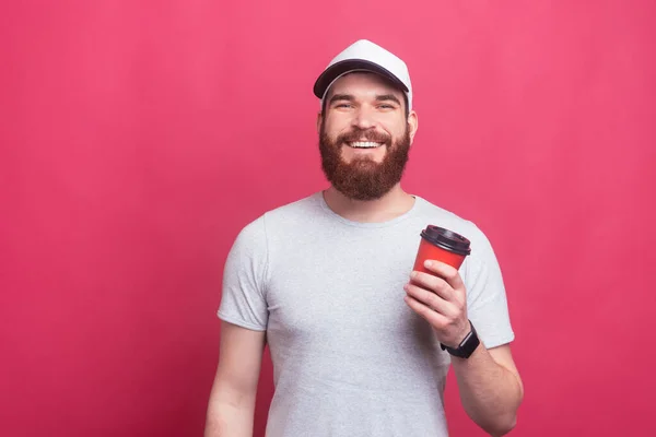 Schöner junger Mann mit Schirmmütze, der eine Tasse Kaffee hält, um über rosa Hintergrund zu gehen — Stockfoto