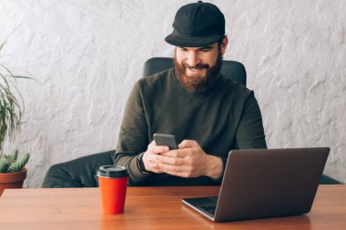 Mutlu genç sakallı adam akıllı telefonunu kullanıp ofisinde oturuyor.