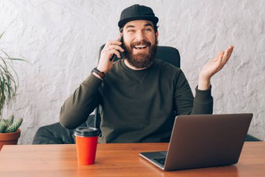 Ofiste otururken akıllı telefondan konuşan yakışıklı, neşeli genç adam.