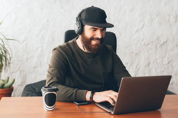 Joven con la barba sonriendo y usando auriculares y trabajando en el ordenador portátil en la oficina —  Fotos de Stock