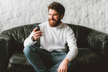 Mutlu genç adam akıllı telefonunu kullanıp koltukta oturuyor.