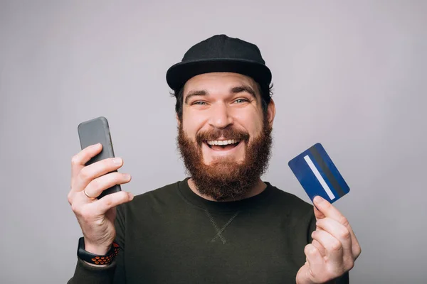 Un joven sonriente sosteniendo una tarjeta y un teléfono. Compras y pagos en línea . — Foto de Stock
