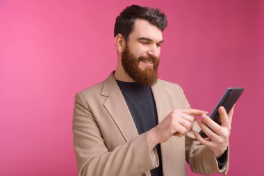 Neşeli genç sakallı adam tabletini pembe arka planda kullanıyor..