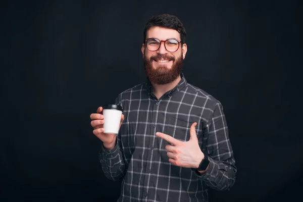 Schöner bärtiger Mann zeigt auf seinen Take-away-Becher auf schwarzem Hintergrund. — Stockfoto