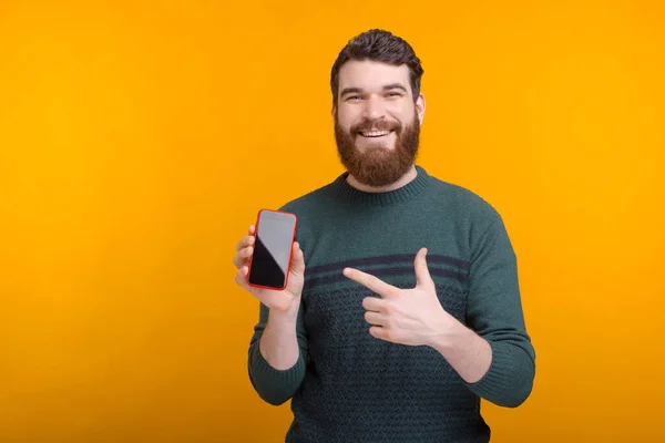 Un homme heureux portant la barbe tient son téléphone et le pointe du doigt . — Photo