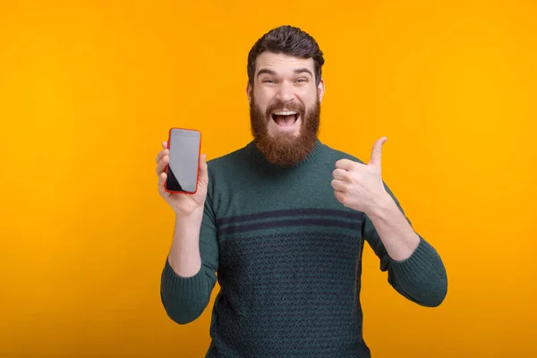 Homem barbudo saiu está olhando para a câmera mostrando o polegar para cima e sua tela do telefone . — Fotografia de Stock