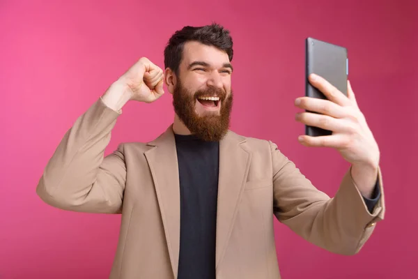 Homem barbudo animado está olhando para seu tablet ou fazendo uma selfie enquanto mostra o gesto vencedor . — Fotografia de Stock