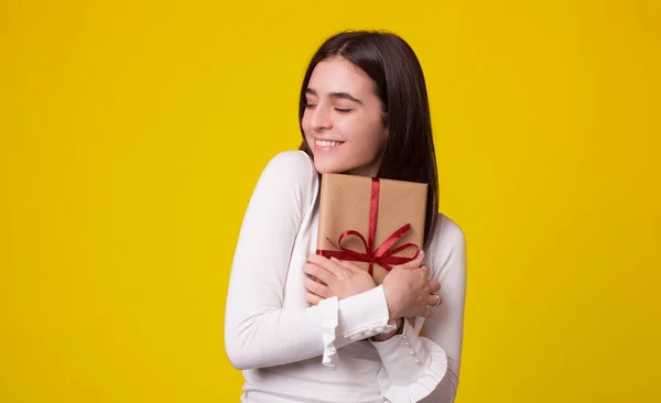 Nettes Mädchen umarmt ein verpacktes Geschenk auf gelbem Hintergrund. — Stockfoto