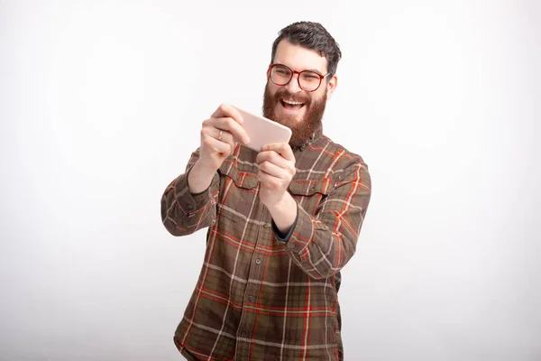 Barbudo hombre está disfrutando jugando con su teléfono sobre fondo blanco . — Foto de Stock