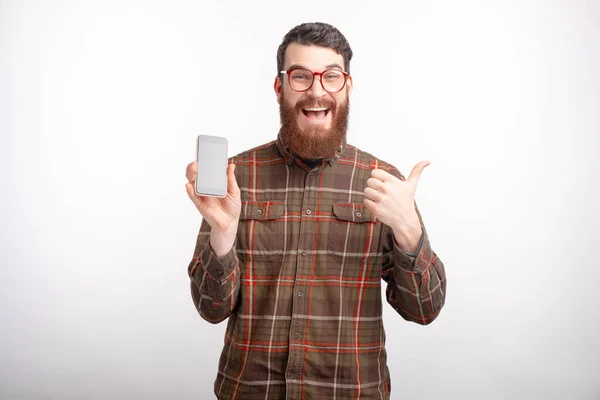Hombre barbudo excitado está mostrando como gesto y su teléfono sobre fondo blanco . — Foto de Stock