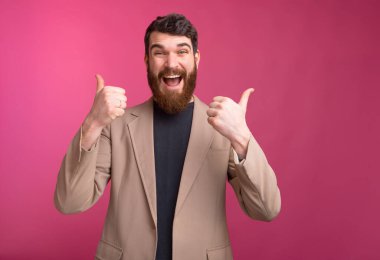 Büyük gülümsemeli, ceketli, sakallı adam pembe arka planda iki parmağını da gösteriyor..