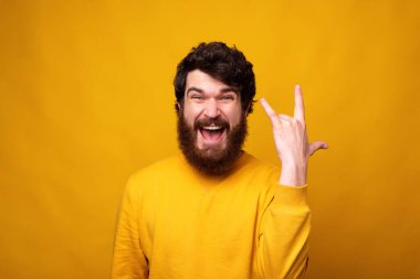 Heyecanlı sakallı adam sarı arka planda kaya jesti yapıyor..