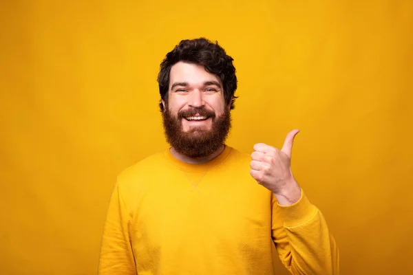 Bello uomo barbuto sta mostrando come o pollice su sfondo giallo . — Foto Stock