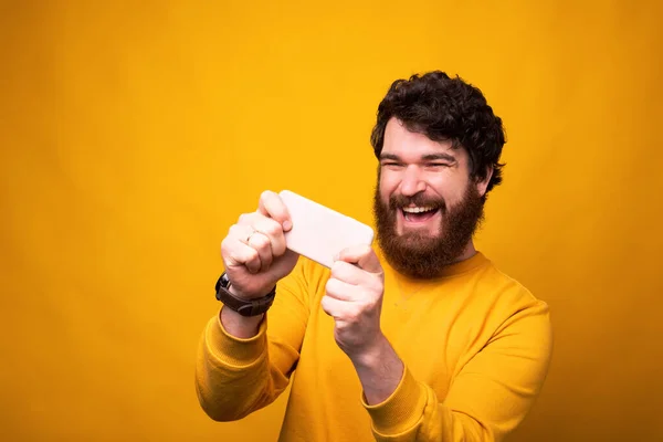 喜怒无常的大胡子男人在黄色背景的手机上玩耍. — 图库照片