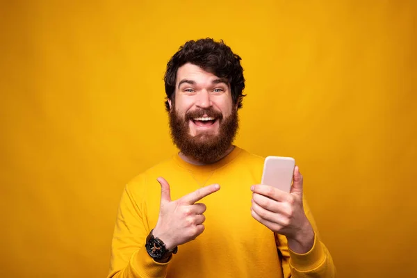 Feliz hombre barbudo está apuntando a su teléfono que está sosteniendo en el fondo amarillo . — Foto de Stock