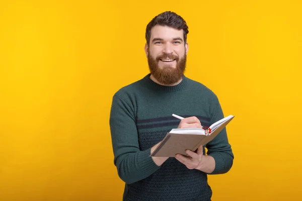 Bärtiger Mann blickt in die Kamera und schreibt irgendetwas ins Tagebuch. — Stockfoto