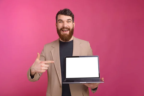 Hombre barbudo excitado está apuntando a su portátil que está sosteniendo en el fondo rosa . — Foto de Stock