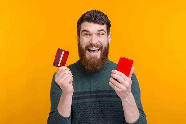 Aufgeregt zeigt ein bärtiger Mann seine Karte und sein Handy. Glücklich über mobiles Banking. — Stockfoto