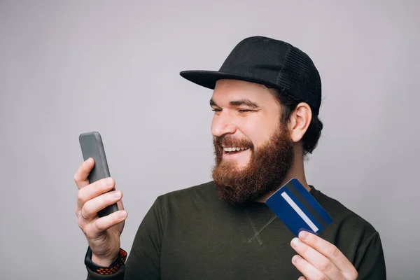 El hombre barbudo disfruta usando la banca móvil. Él sostiene su teléfono y su tarjeta . — Foto de Stock