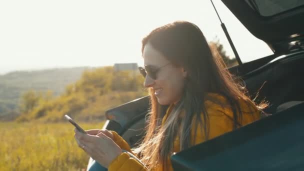 Ung kvinna reser med bil och använder sin smartphone under solnedgången — Stockvideo