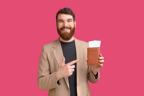 Усміхнений бородатий чоловік вказує на свій паспорт і квитки, які він тримає на рожевому тлі . — стокове фото
