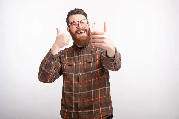 Sonriendo barbudo hombre está mostrando el pulgar hacia arriba está haciendo una selfie sobre blanco . —  Fotos de Stock