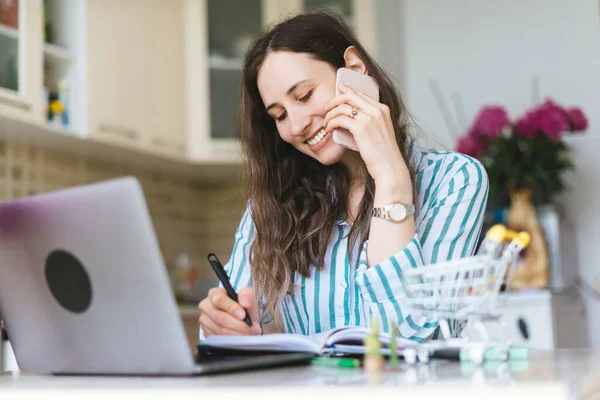 Gülümseyen genç bir kadın evde çalışıyor ve akıllı telefondan konuşuyor. — Stok fotoğraf