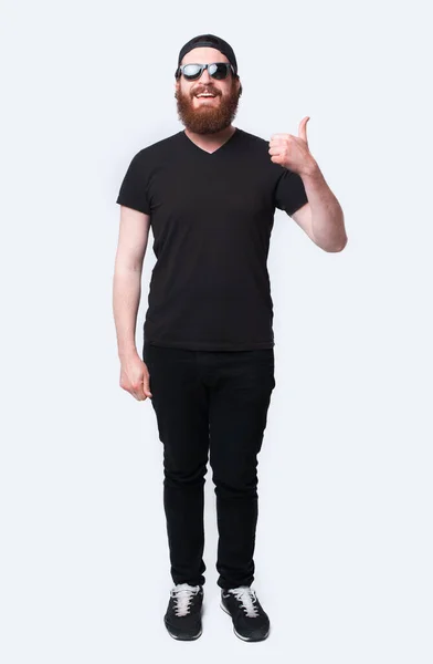Foto de comprimento total do homem hipster barbudo usando óculos de sol e mostrando o polegar sobre o branco — Fotografia de Stock