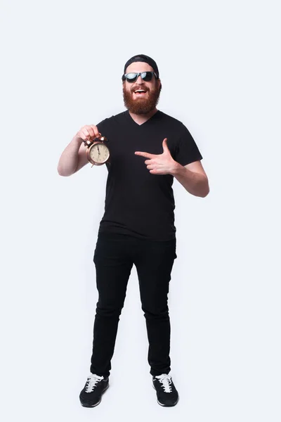 Full length photo smiling bearded man pointing at alarm clock over white — Stock Photo, Image