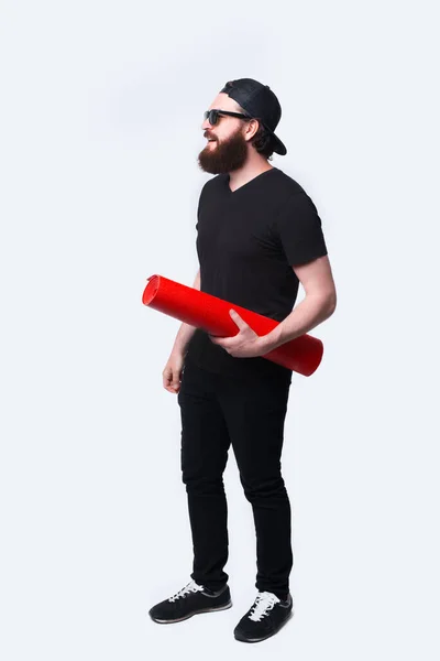 Pełna długość zdjęcie wesołego brodatego mężczyzny z czerwoną matą do jogi stojącego nad białym — Zdjęcie stockowe